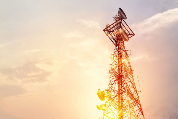 A tall communication tower with multiple antennas stands against a setting sun.
