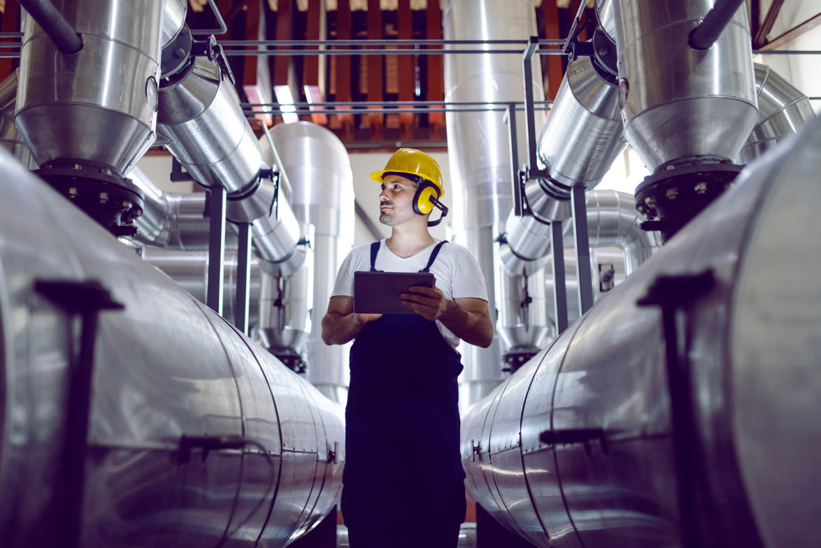 A worker in a hard hat and ear protection inspects a brewery, holding a tablet and standing between large stainless steel tanks.