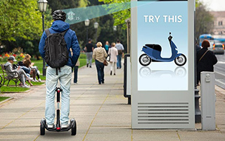 A person riding a Segway looks at a digital advertisement board displaying a blue scooter with the text "TRY THIS" in an urban park setting.