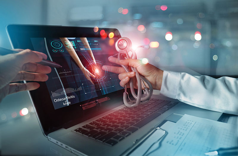 A doctor, utilizing 5G in connected healthcare, points to a virtual screen displaying a knee with data, while holding a stethoscope. Another hand with a pen appears on the left. The laptop showcases medical imagery and charts, highlighting cutting-edge technology in modern medicine.