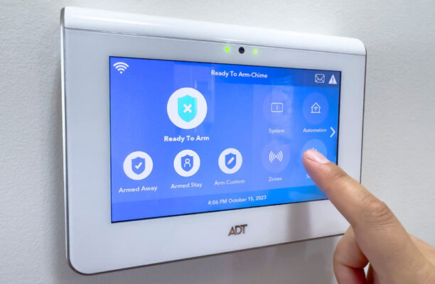 A finger touches a home security system display, navigating options like "Armed Away," "Armed Stay" and "Zones." The screen indicates it's "Ready to Arm."