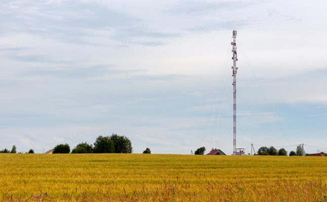 A tall communication tower stands in a field of golden wheat, with a few scattered trees and a small house in the background under a cloudy sky.